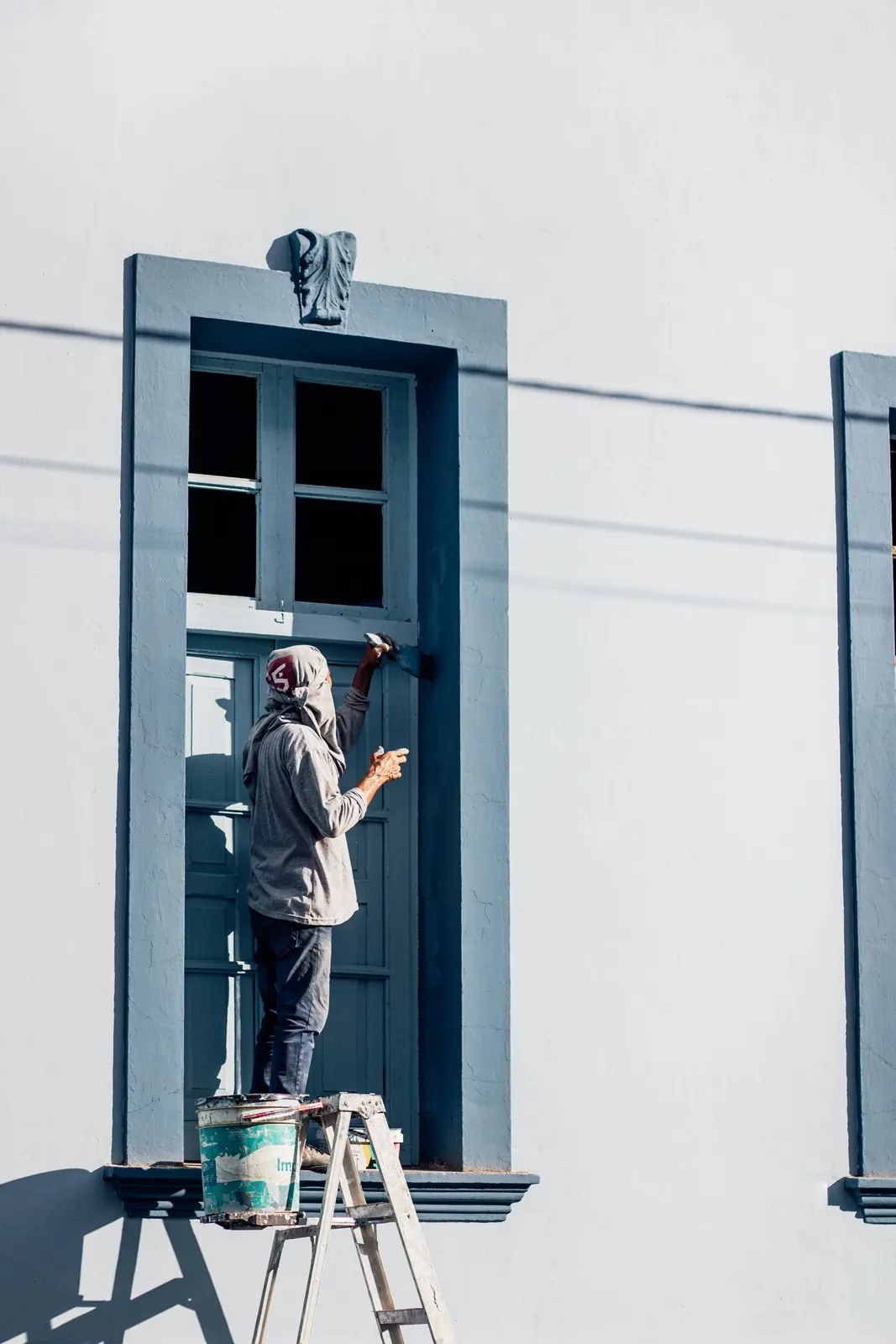 A painter on a ladder brushing a dark blue window frame on the white outside wall of a building