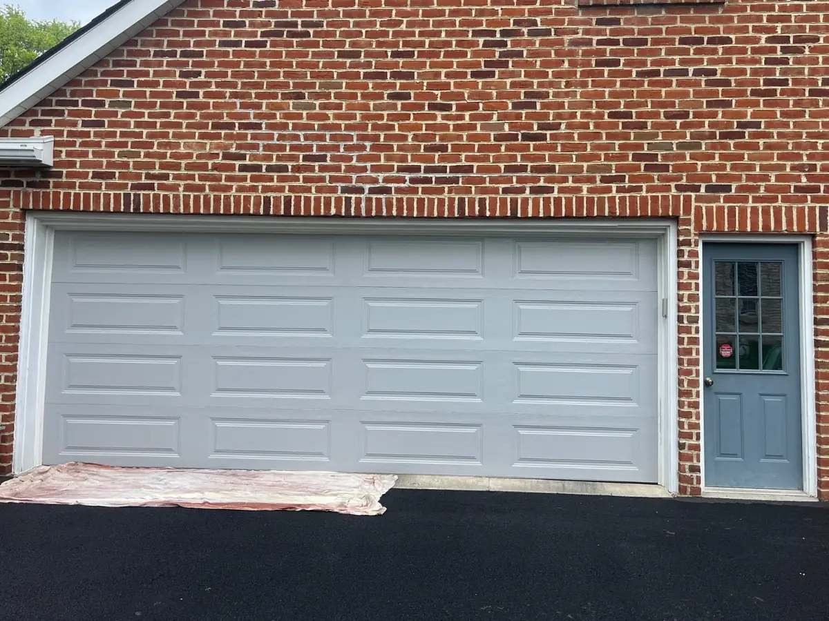 Double garage door before painting, light gray, in a red brick wall, with a drop cloth on the driveway
