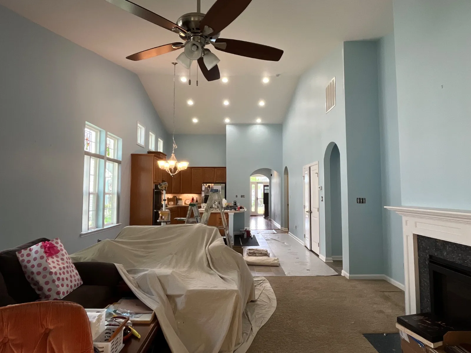 Living room with light blue walls mid-job, the furniture under drop cloths and two stepladders by the kitchen