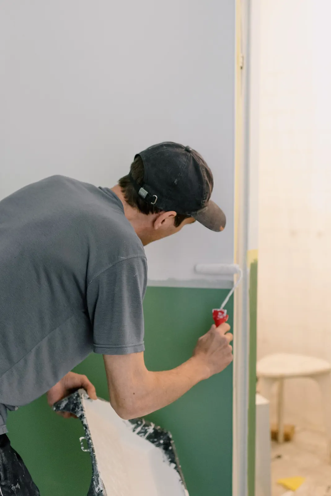 A painter in a gray t-shirt and cap rolling a green wall where it meets a white one in an emptied room