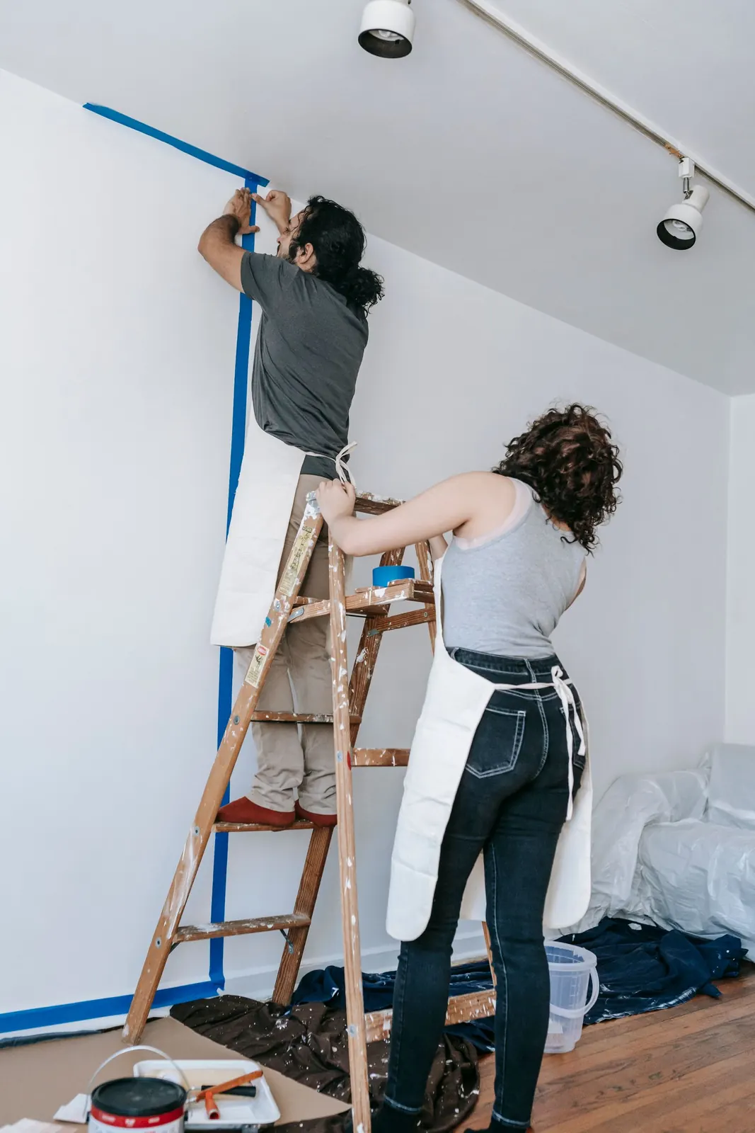 A painter on a wooden stepladder running masking tape along the line between wall and ceiling